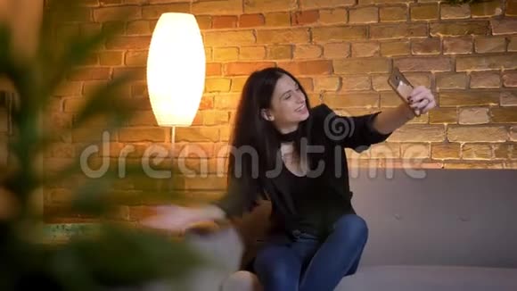 年轻的黑发白种人女孩坐在沙发上在家庭氛围中使用智能手机在视频聊天的肖像视频的预览图