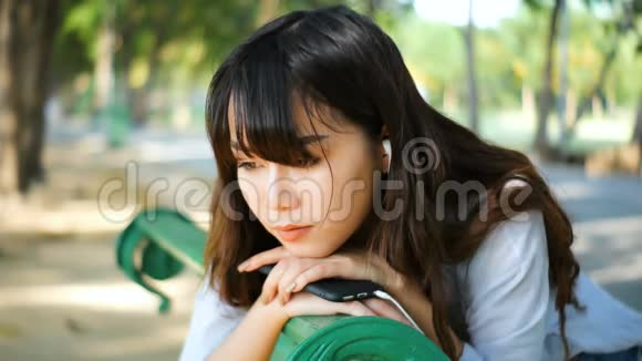美丽的年轻时髦女郎悲伤的亚洲女人带着智能手机带着耳机听音乐坐在公园的长凳上视频的预览图
