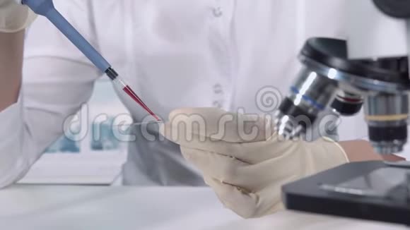 戴手套的手的特写镜头一位女性研究人员在显微镜下用微量吸管和玻璃进行血液测试视频的预览图