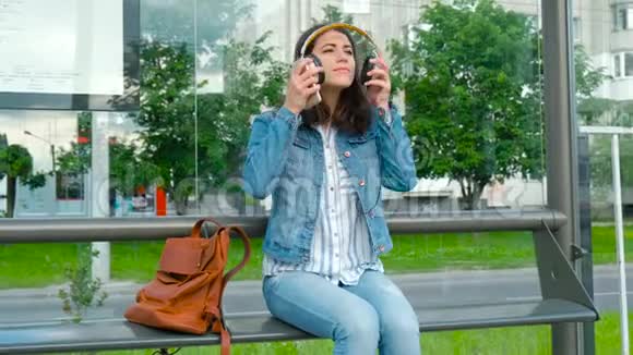 年轻女子戴着耳机欣赏音乐坐在现代有轨电车车站等公共交通视频的预览图