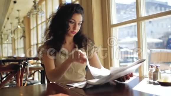一个卷发女人坐在咖啡馆的窗户附近看报纸卷发女人坐在咖啡馆和树叶的窗户附近视频的预览图