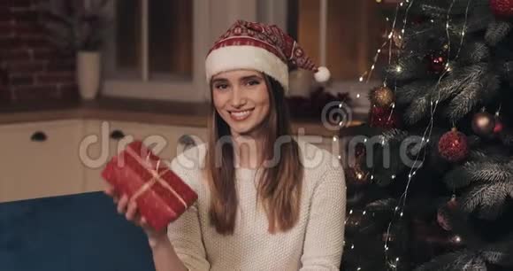 在舒适的圣诞节年轻漂亮的女人戴着桑塔帽坐在圣诞树附近的沙发上的肖像视频的预览图