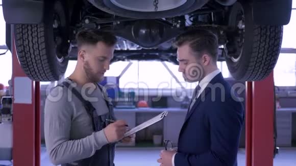 汽车服务机械师建议客户并在车下的剪贴板上做笔记然后男人握手和视频的预览图