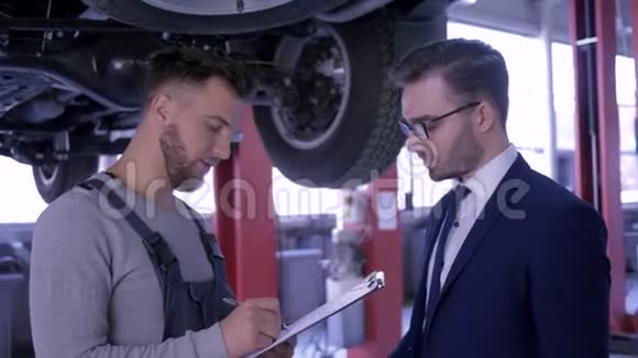 机械师为顾客提供建议并在汽车升降台上的固定板上做笔记视频的预览图