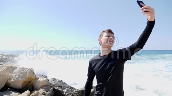 年轻的花式运动健康的白种人穿着黑色衣服站在岩石海滩上时一边自拍一边微笑一边海浪拍打和哭泣视频的预览图