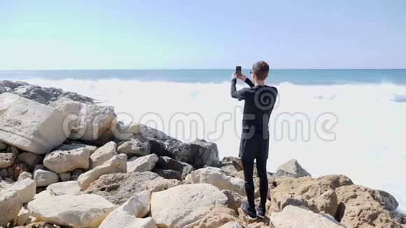 一个年轻的白种人站在岩石海滩上手里拿着电话一边说着话一边拍着海浪拍打岩石的照片视频的预览图