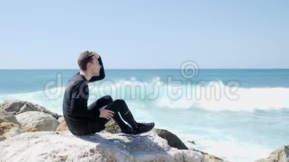 年轻的白种人坐在岩石上手指插在头发上强烈的海浪拍打着溅起水花的岩石海滩视频的预览图