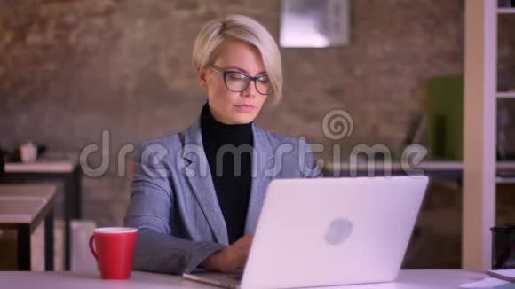 金发短发女商人戴着眼镜看着笔记本电脑屏幕和在办公室喝咖啡的肖像视频的预览图