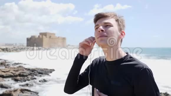 年轻的白种人把耳机放在岩石海滩上背景是城堡视频的预览图