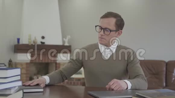 年轻谦逊的戴着眼镜的男人坐在办公室的木桌旁许多书都在桌子上帅哥视频的预览图