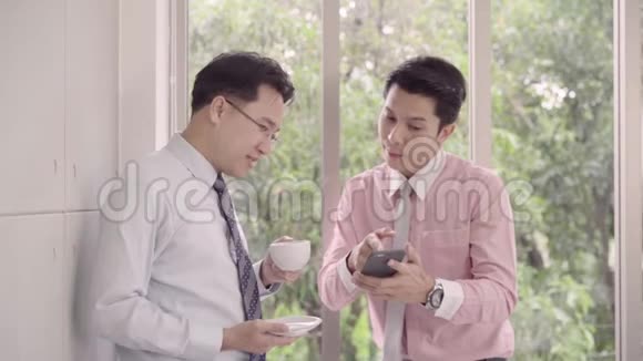 慢动作年轻人微笑着一边站着喝热咖啡一边在办公室放松使用智能手机的亚洲商人视频的预览图