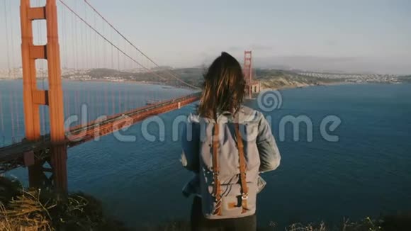 背着背包的年轻女子旅行者头发随风飘扬在金门大桥欣赏风景如画的日落视频的预览图