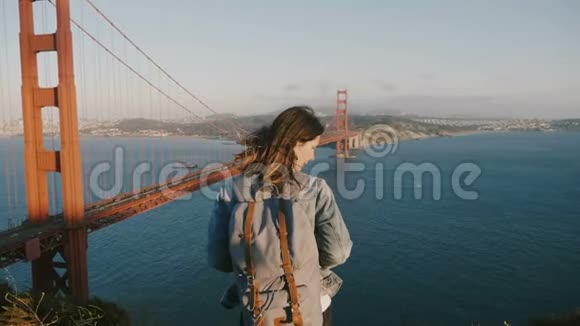 镜头放大年轻幸福的女子背后背包头发在风中欣赏日落风景在金门大桥视频的预览图