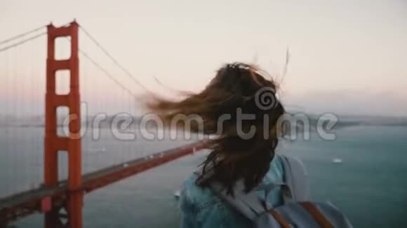 在标志性的金门大桥景观中年轻的快乐旅游妇女带着背包在她的头发着强烈的风视频的预览图