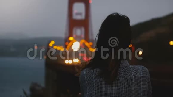后景年轻女子独自站在深夜观看标志性的金门大桥交通视图下的大风视频的预览图