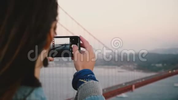 后景特写年轻女士拍摄智能手机照片惊人的日落金门大桥在旧金山美国视频的预览图