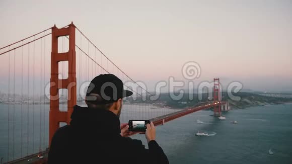后景年轻兴奋的美国男人穿着休闲服装拍摄智能手机照片日落金门大桥加利福尼亚州视频的预览图