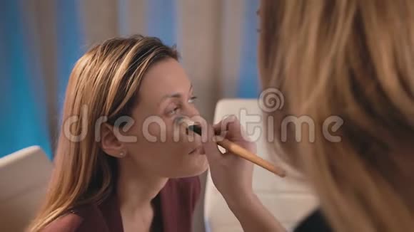 一个化妆师在一个白人金发模特的脸上涂了一把刷子化妆生意的女人家庭视频的预览图