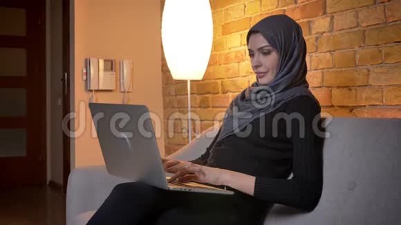 戴头巾的穆斯林成年女性的特写照片她坐在室内沙发上手提电脑视频的预览图