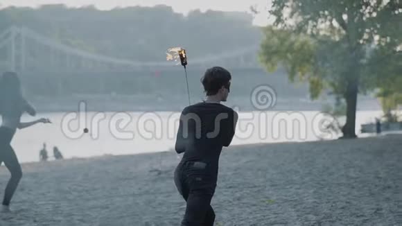 年轻苗条的男人穿着黑色的衣服女人站在河岸上用火焰表演节目熟练的射击技巧视频的预览图