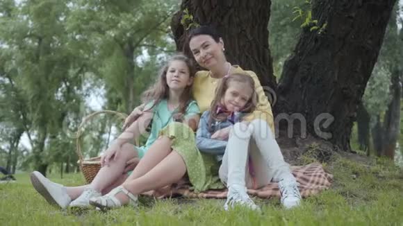 肖像高级妇女亲吻和拥抱可爱的孙女坐在公园树下的草地上看着视频的预览图