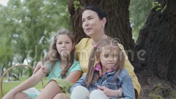 肖像成熟可爱的女人亲吻拥抱可爱的孙女坐在公园树下的草地上视频的预览图