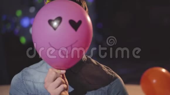 一个自信的年轻人戴着生日帽从头上摘下一个粉红色的气球上面画着一张滑稽的脸视频的预览图