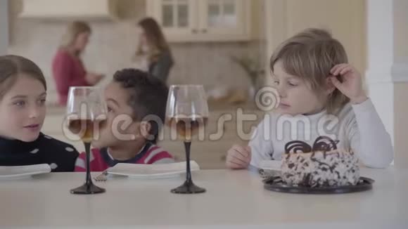 三个有趣的孩子坐在桌子旁拿着小蛋糕和带果汁的酒杯两个高加索女孩和非洲女孩视频的预览图