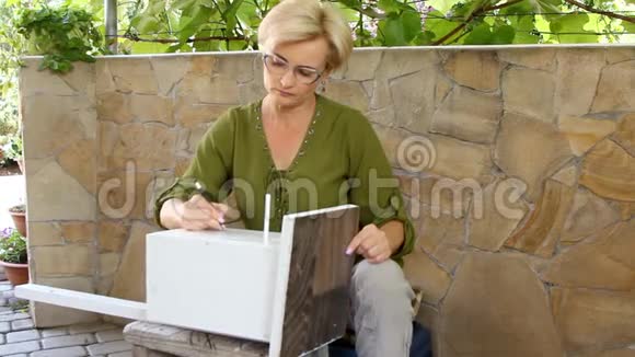 美丽的女人在一个木制的筑巢箱上画着一只绿鸟的轮廓它与葡萄的绿叶交织在一起视频的预览图