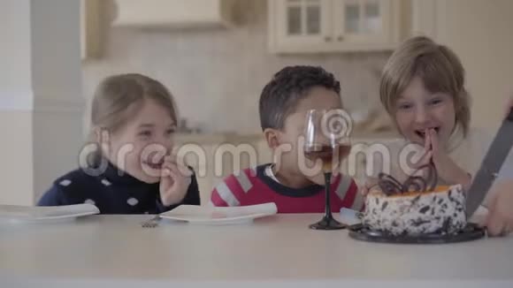 三个孩子在桌子上拿着小蛋糕和果汁眼镜女人的手为孩子切蛋糕两名白种人视频的预览图