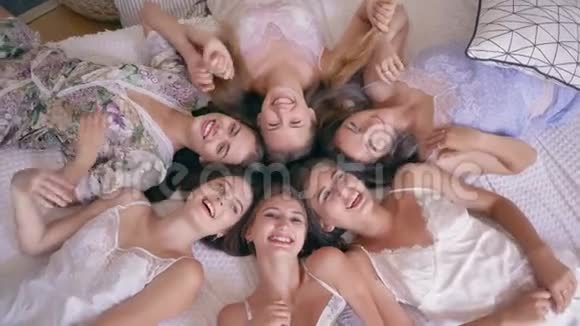 睡衣派对六个穿着时髦睡衣微笑的年轻女性躺在床上在房间里直视镜头视频的预览图