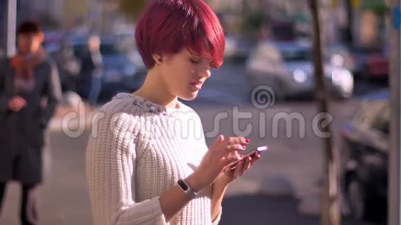 忙碌的粉红色头发女孩的肖像带着智能手机转向相机在模糊的街道背景上微笑视频的预览图