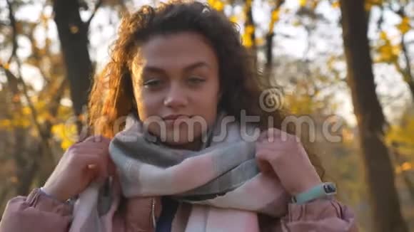 在秋日公园里一位白种人卷发女子在镜头前观看并藏在温暖的围巾中的特写镜头视频的预览图