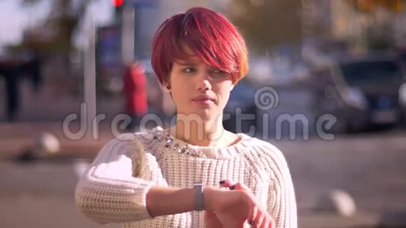 在模糊的街道背景下用手表打电话的快乐漂亮的粉红色头发女孩的肖像视频的预览图