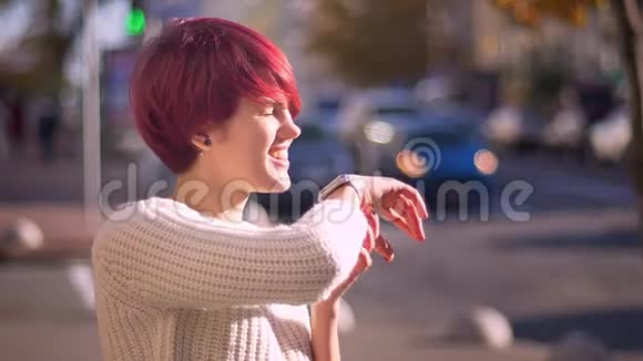 在模糊的街道背景下用手表打电话给一个情绪激动的漂亮粉红头发女孩的肖像视频的预览图