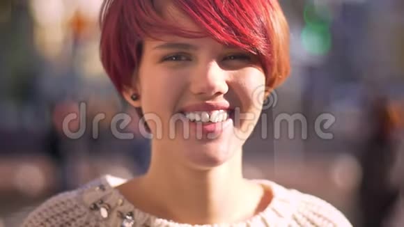 在模糊的街道背景下年轻的白种人粉红头发女孩谦卑地微笑着进入相机的特写肖像视频的预览图