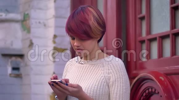 年轻的白种人粉红头发女孩的肖像愉快地与智能手机在红色的门背景下工作视频的预览图