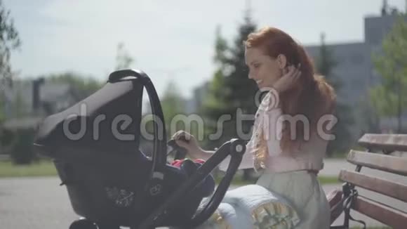 在公园的特写镜头中美丽的红发女人和她的孩子躺在婴儿车里玩耍她很享受视频的预览图