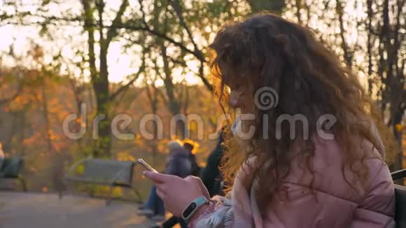 白种人卷发女子坐在长凳上在秋天快乐地看着智能手机和向上的画像视频的预览图