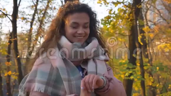 白种人卷发女人看着她的手表在秋天公园的背景上转向相机的肖像视频的预览图