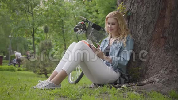 肖像可爱的微笑的女人坐在公园的一棵老树下大声朗读日记现代自行车站着视频的预览图