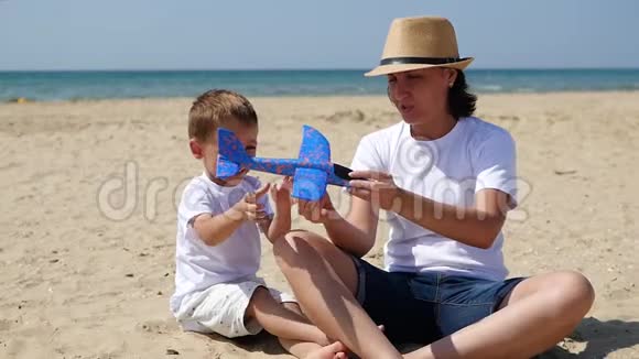 一个微笑的小男孩玩飞机描绘飞行宝宝和妈妈正在休息坐在沙滩上视频的预览图