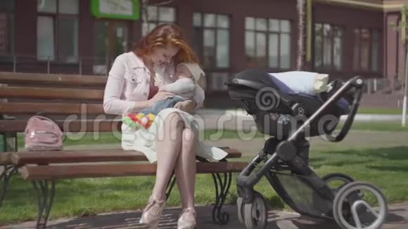 美丽的红发女人和坐在长凳上的孩子玩站在附近的婴儿车她很享受视频的预览图