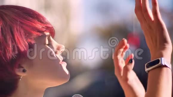 年轻的白种人粉红色头发的女孩的特写肖像微笑着通过她的手在阳光下观看视频的预览图