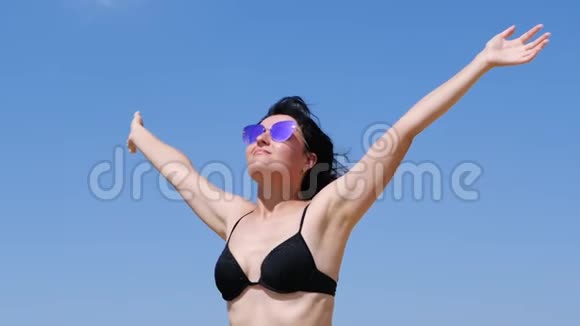 一个年轻的黑发女孩站在海滨双臂举向天空一个女人举起双臂对着蓝色视频的预览图