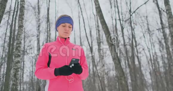 一位正在晨跑的年轻女性手里拿着一部智能手机选择一条音乐曲目进行训练冬季跑步使用视频的预览图