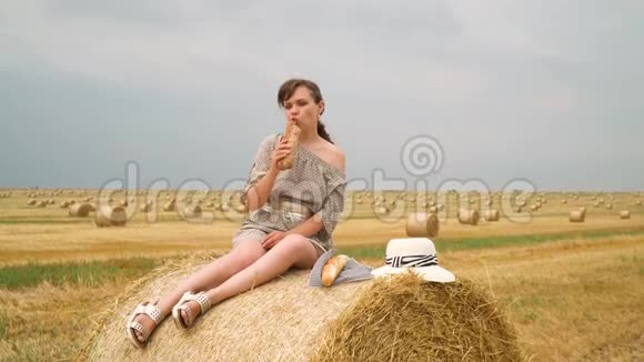 一个年轻的女孩坐在蓓蕾的顶上夏天的晚上在麦田中央吃面包视频的预览图