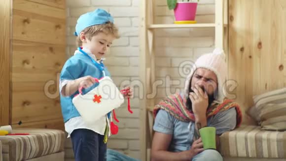 男孩和父亲扮演医生和病人那个穿医生西装男孩用针织帽子招待一个留胡子的人概念视频的预览图