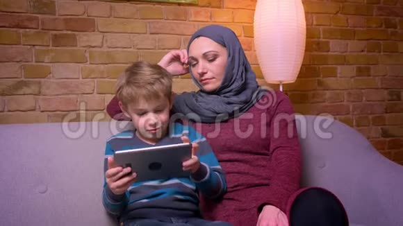 一个固执的小男孩在平板电脑上玩游戏他的穆斯林母亲戴着头巾在家里观察他的活动视频的预览图