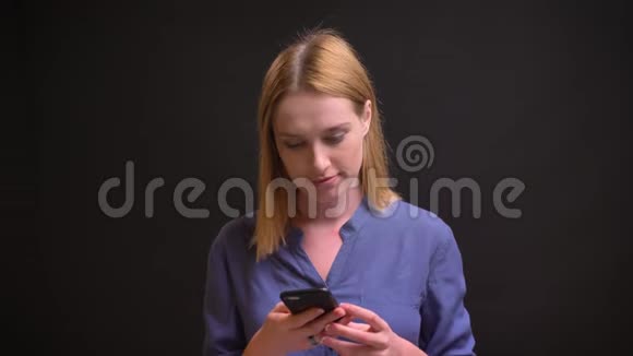 白种人金发女人穿着衬衫在黑色背景下专注地看着智能手机的肖像视频的预览图